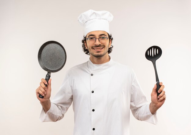 Sorridente giovane cuoco maschio che indossa l'uniforme del cuoco unico e bicchieri che tengono padella e spatola isolato sulla parete bianca