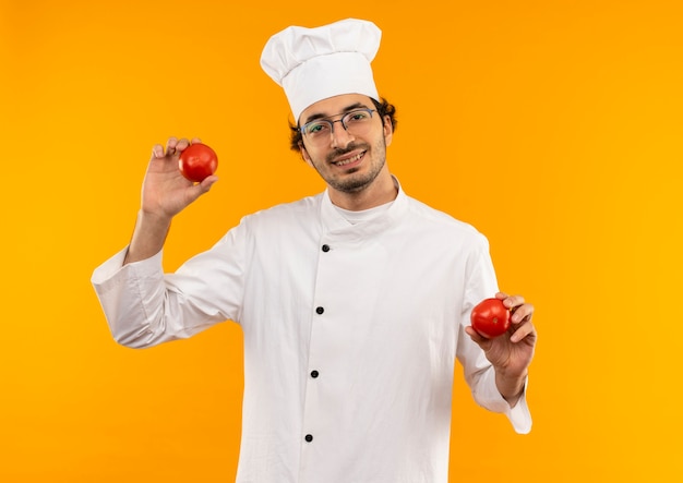 Sorridente giovane cuoco maschio che indossa l'uniforme del cuoco unico e bicchieri che tengono pomodoro isolato sulla parete gialla