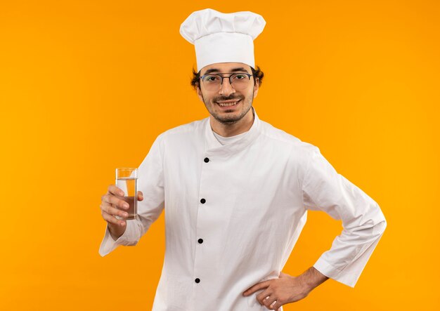 Sorridente giovane cuoco maschio che indossa l'uniforme dello chef e bicchieri tenendo il bicchiere d'acqua e mettendo la mano sul fianco isolato sulla parete gialla