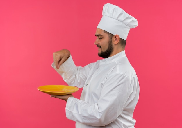 Sorridente giovane cuoco maschio in uniforme chef tenendo piatto e aggiunta di sale isolato su spazio rosa