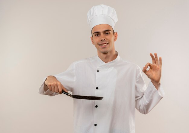 Sorridente giovane cuoco maschio indossa uniforme del cuoco unico che tiene padella che mostra okey gesto