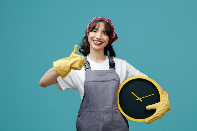 Sorridente giovane donna addetta alle pulizie che indossa bandana uniforme e guanti di gomma che tengono l'orologio guardando la fotocamera che mostra il pollice in su isolato su sfondo blu