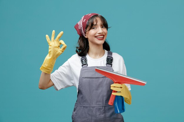 Sorridente giovane donna addetta alle pulizie che indossa bandana uniforme e guanti di gomma che tengono tergicristallo e detergente guardando la fotocamera che mostra segno ok isolato su sfondo blu