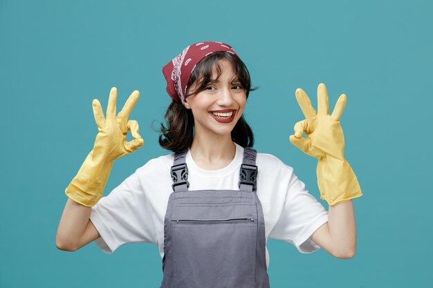 sorridente giovane donna addetta alle pulizie che indossa bandana uniforme e guanti di gomma guardando la fotocamera che mostra il segno ok con entrambe le mani isolate su sfondo blu