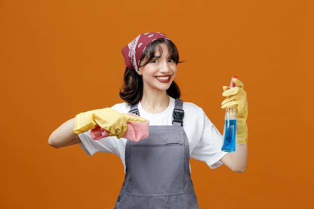 Sorridente giovane donna addetta alle pulizie che indossa guanti di gomma uniformi e bandana che tiene panno per spolverare e detergente guardando la fotocamera che punta al detergente isolato su sfondo arancione