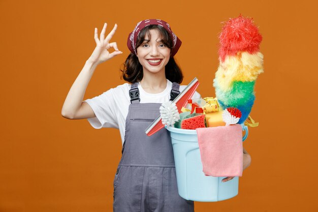 Sorridente giovane donna addetta alle pulizie che indossa uniforme e bandana con secchio di strumenti per la pulizia guardando la fotocamera che mostra il segno ok isolato su sfondo arancione