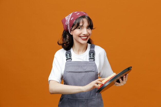 Sorridente giovane donna addetta alle pulizie che indossa uniforme e bandana guardando il vassoio per la pulizia della fotocamera con spugna isolata su sfondo arancione