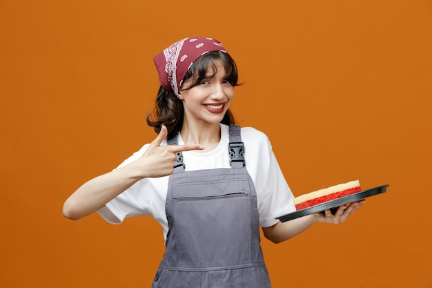 Sorridente giovane donna addetta alle pulizie che indossa uniforme e bandana vassoio con spugna in esso che punta al vassoio guardando la fotocamera isolata su sfondo arancione