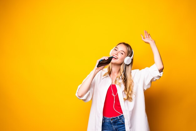 Sorridente giovane donna ascoltando musica in cuffia e utilizzando smartphone isolato sopra la parete gialla