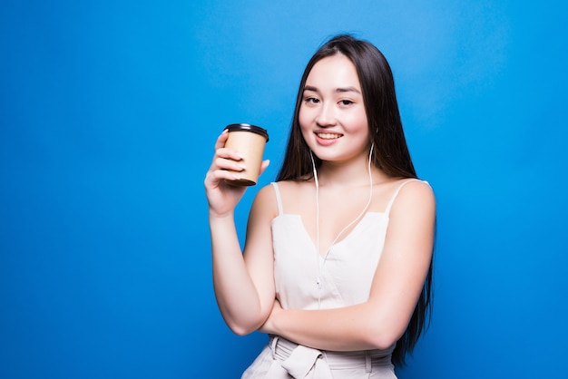 Sorridente giovane donna asiatica che tiene tazza di caffè da asporto in piedi isolato sopra la parete blu