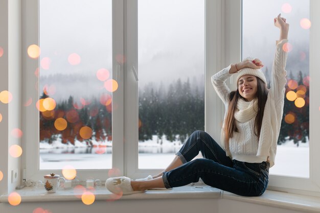 Sorridente giovane donna attraente felice in elegante maglione bianco lavorato a maglia, sciarpa e cappello seduto a casa sul davanzale della finestra a Natale divertendosi alzando le mani, vista di sfondo foresta invernale, luci bokeh