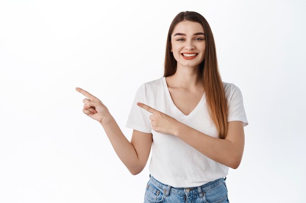 Sorridente giovane donna attraente presenta banner, indicando da parte l'offerta promozionale del logo, sembra soddisfatta, consiglia il prodotto, in piedi in t-shirt contro il muro bianco