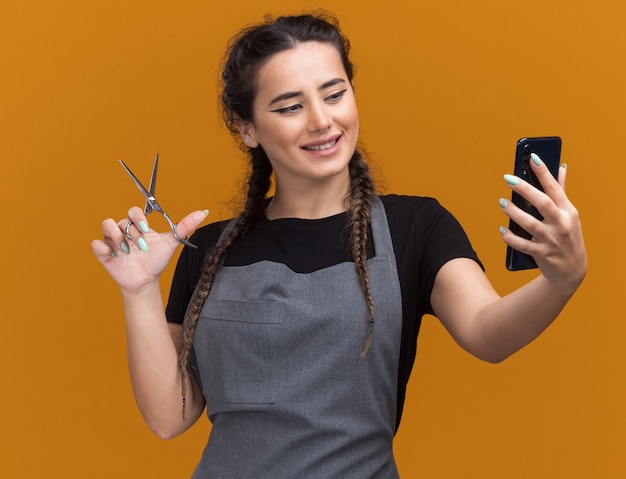 Sorridente giovane donna barbiere in uniforme che tiene le forbici e guardando il telefono in mano isolato sulla parete arancione