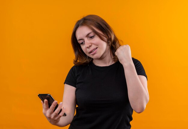 Sorridente giovane donna casual tenendo il telefono cellulare e guardandolo con il pugno alzato su uno spazio arancione isolato