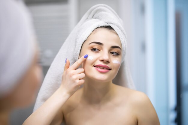 Sorridente giovane donna che applica la crema sul viso e cerca di specchiarsi nel bagno di casa