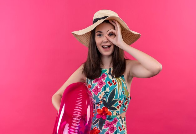 Sorridente giovane donna che indossa il cappello che tiene l'anello di nuotata e che fa segno giusto sulla parete rosa isolata