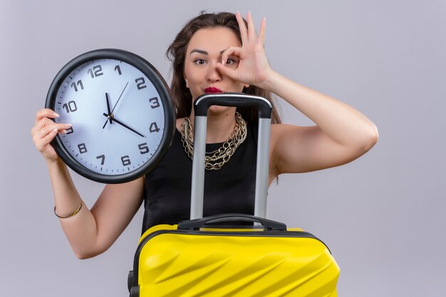 Sorridente giovane donna che indossa la maglietta nera che tiene l'orologio da parete e mostra il gesto okey sulla parete bianca