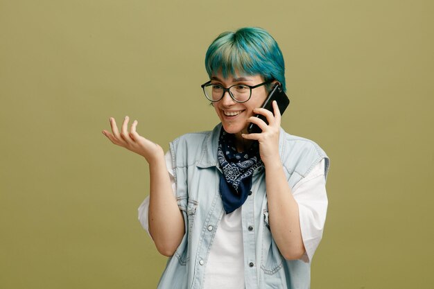 Sorridente giovane donna che indossa occhiali bandana sul collo guardando il lato parlando al telefono che mostra la mano vuota isolata su sfondo verde oliva