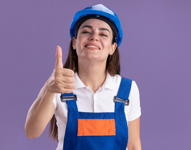 Sorridente giovane donna costruttore in uniforme che mostra pollice in alto isolato sulla parete viola