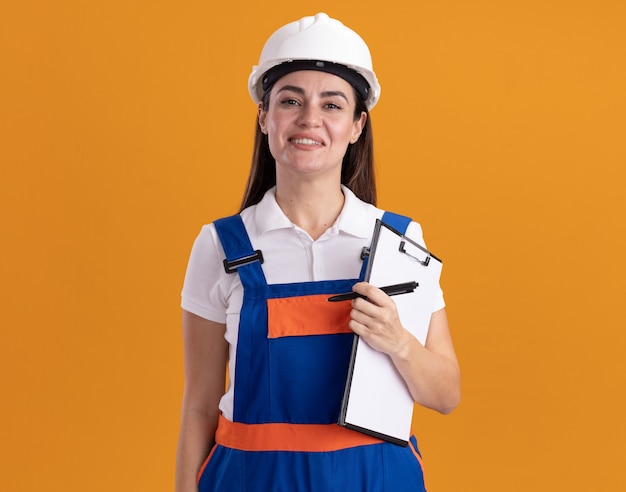 Sorridente giovane donna costruttore in uniforme che tiene appunti con la penna isolata sulla parete arancione