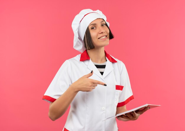 Sorridente giovane donna cuoco in uniforme del cuoco unico che tiene e che indica al blocco note isolato sulla parete rosa