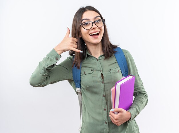 Sorridente giovane donna della scuola con gli occhiali con zaino in possesso di libri che mostrano il gesto della telefonata