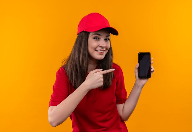 Sorridente giovane donna delle consegne che indossa la maglietta rossa in berretto rosso che tiene il telefono da un lato e indica con l'altra mano sul muro giallo isolato