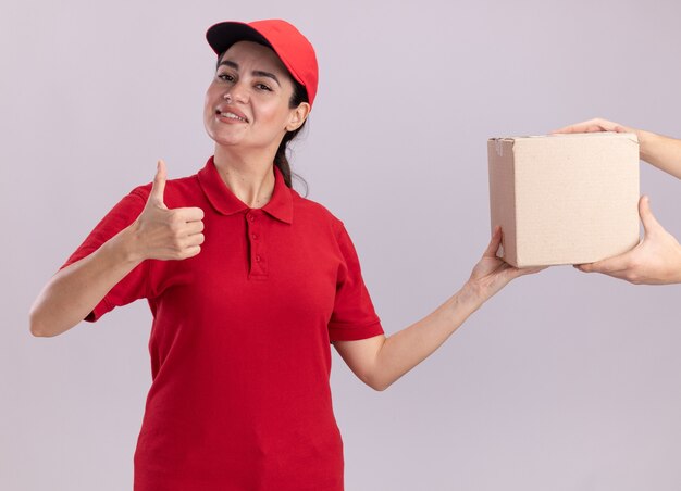 Sorridente giovane donna delle consegne in uniforme e berretto che dà cardbox al cliente che mostra pollice in su