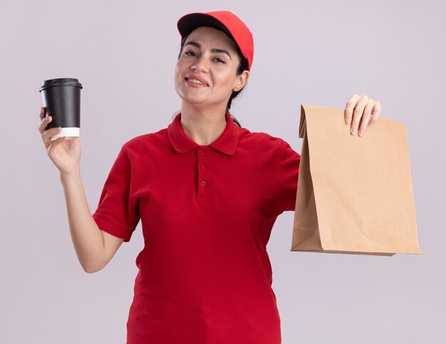 Sorridente giovane donna delle consegne in uniforme e berretto che tiene una tazza di caffè in plastica e un pacchetto di carta