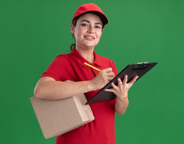 Sorridente giovane donna delle consegne in uniforme e berretto con scatola di cartone e appunti con matita