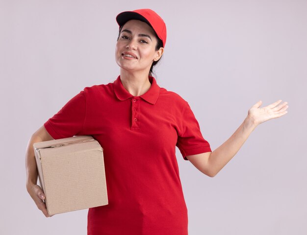 Sorridente giovane donna delle consegne in uniforme e cappuccio che tiene una scatola di cartone che mostra la mano vuota