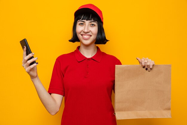 Sorridente giovane donna di consegna caucasica che tiene in mano un sacchetto di carta per alimenti e un telefono