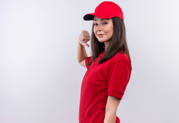 Sorridente giovane donna di consegna che indossa la maglietta rossa in berretto rosso che mostra i pollici verso il basso sulla parete bianca isolata