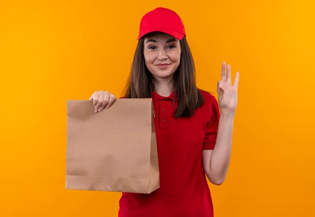 Sorridente giovane donna di consegna che indossa la maglietta rossa in berretto rosso che tiene il pacchetto e mostra il gesto okey sulla parete gialla isolata