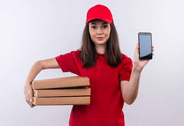 Sorridente giovane donna di consegna che indossa la maglietta rossa in berretto rosso che tiene la scatola della pizza e telefono sul muro bianco isolato