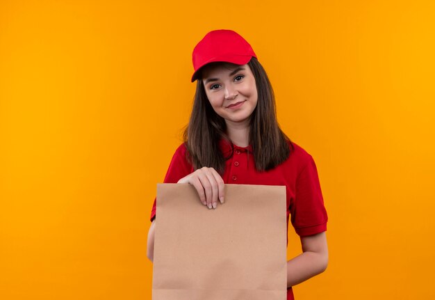 Sorridente giovane donna di consegna che indossa la maglietta rossa in berretto rosso che tiene un pacchetto sulla parete arancione isolata