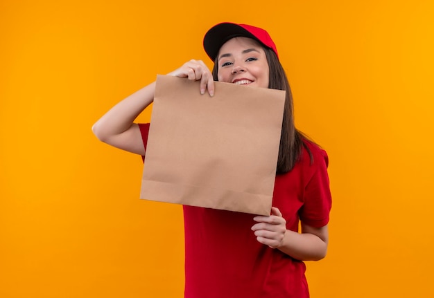 Sorridente giovane donna di consegna che indossa la maglietta rossa in berretto rosso che tiene un pacchetto sulla parete arancione isolata