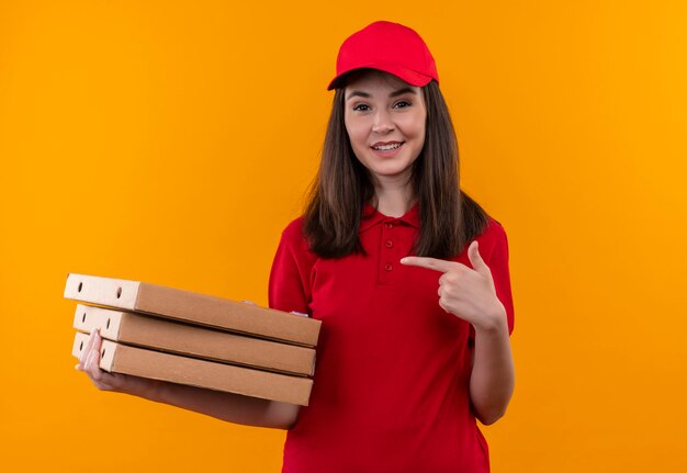 Sorridente giovane donna di consegna che indossa la maglietta rossa in berretto rosso che tiene una scatola di pizza e punti con un dito sulla parete arancione isolata