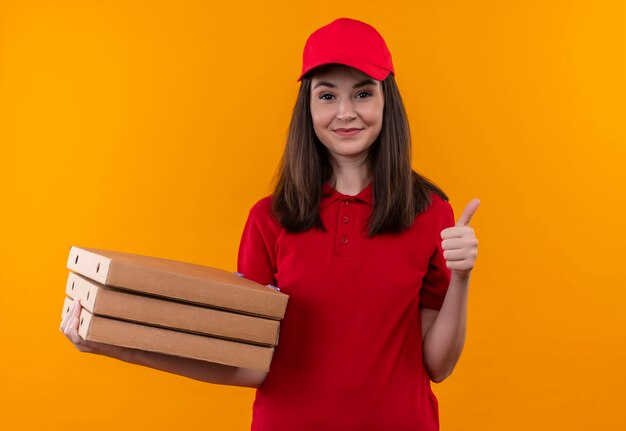 Sorridente giovane donna di consegna che indossa la maglietta rossa in berretto rosso che tiene una scatola di pizza e spettacoli come sulla parete arancione isolata
