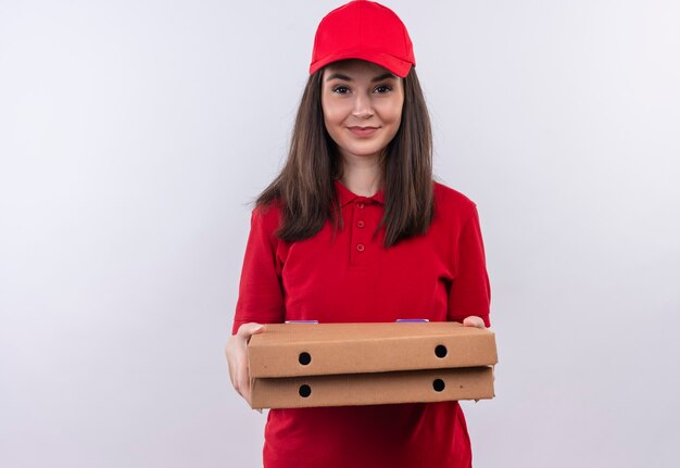 Sorridente giovane donna di consegna che indossa la maglietta rossa in berretto rosso che tiene una scatola di pizza sul muro bianco isolato