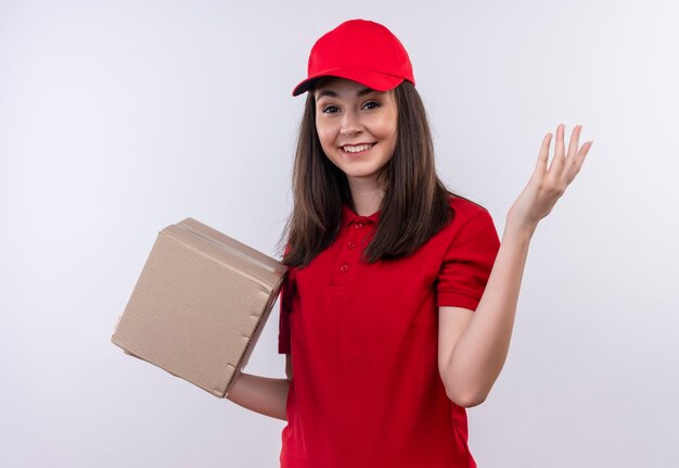 Sorridente giovane donna di consegna che indossa la maglietta rossa in berretto rosso che tiene una scatola e alzò la mano sul muro bianco isolato