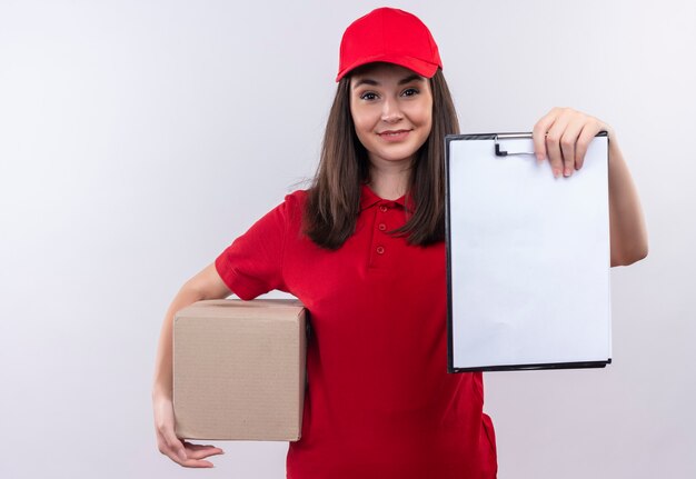 Sorridente giovane donna di consegna che indossa la maglietta rossa in berretto rosso che tiene una scatola e appunti sul muro bianco isolato