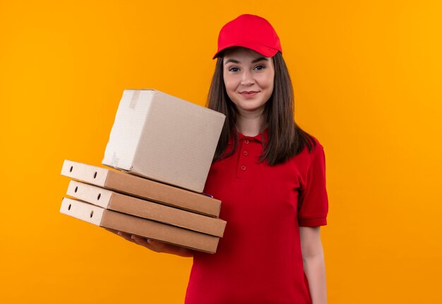 Sorridente giovane donna di consegna che indossa la maglietta rossa in berretto rosso che tiene una scatola e una scatola per pizza sulla parete arancione isolata