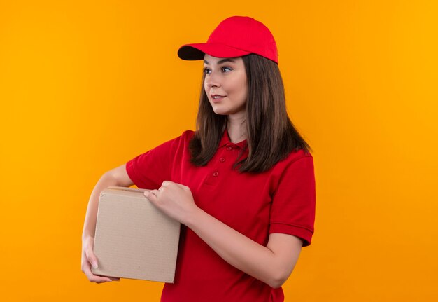 Sorridente giovane donna di consegna che indossa la maglietta rossa in berretto rosso che tiene una scatola sulla parete arancione isolata