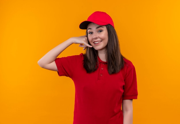 Sorridente giovane donna di consegna che indossa la maglietta rossa in berretto rosso fa una chiamata con le sue mani sulla parete arancione isolata