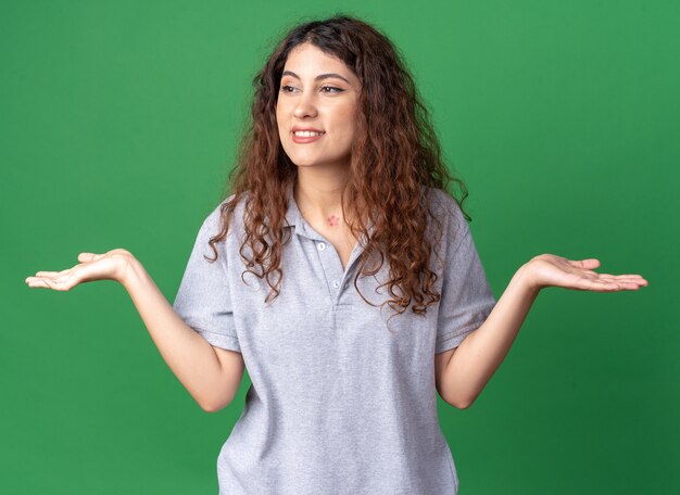 Sorridente giovane donna graziosa che guarda al lato mostrando le mani vuote isolate sul muro verde