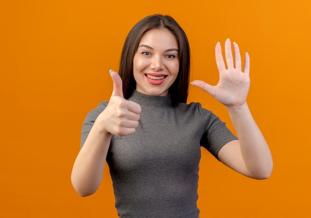 Sorridente giovane donna graziosa che mostra il pollice in alto e cinque con la mano isolata su sfondo arancione