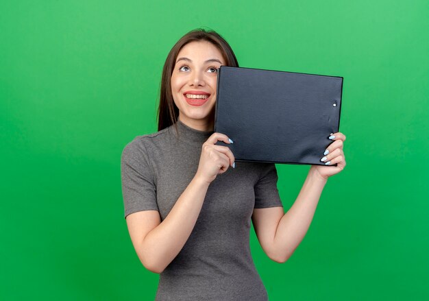 Sorridente giovane donna graziosa che osserva in su tenendo appunti isolato su sfondo verde con spazio di copia