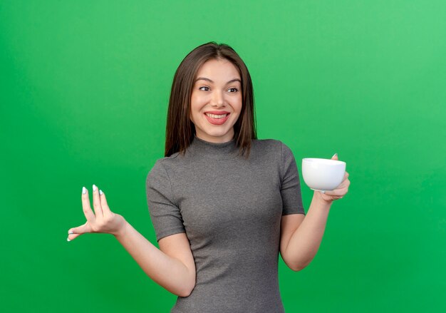 Sorridente giovane donna graziosa che tiene la tazza guardando dritto e tenendo la mano in aria isolato su sfondo verde