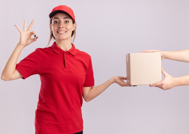 Sorridente giovane donna graziosa di consegna in gesti uniformi segno ok con la mano e dà il cardbox a qualcuno isolato